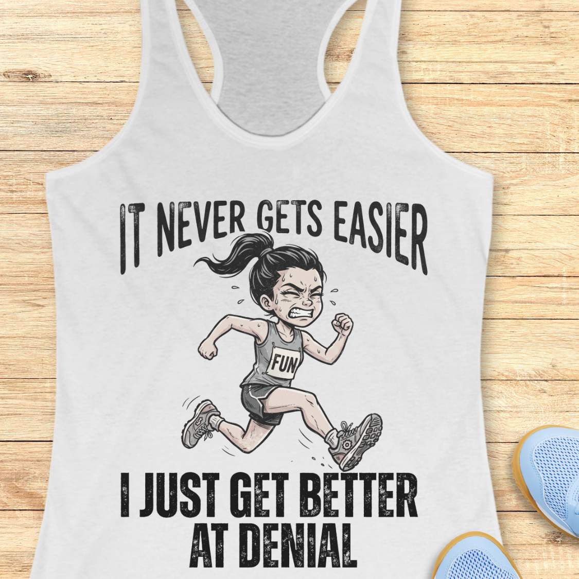 white tank top with black graphic design and text 'it never gets easier, I just get better at denial', on a wooden surface