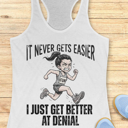white tank top with black graphic design and text 'it never gets easier, I just get better at denial', on a wooden surface