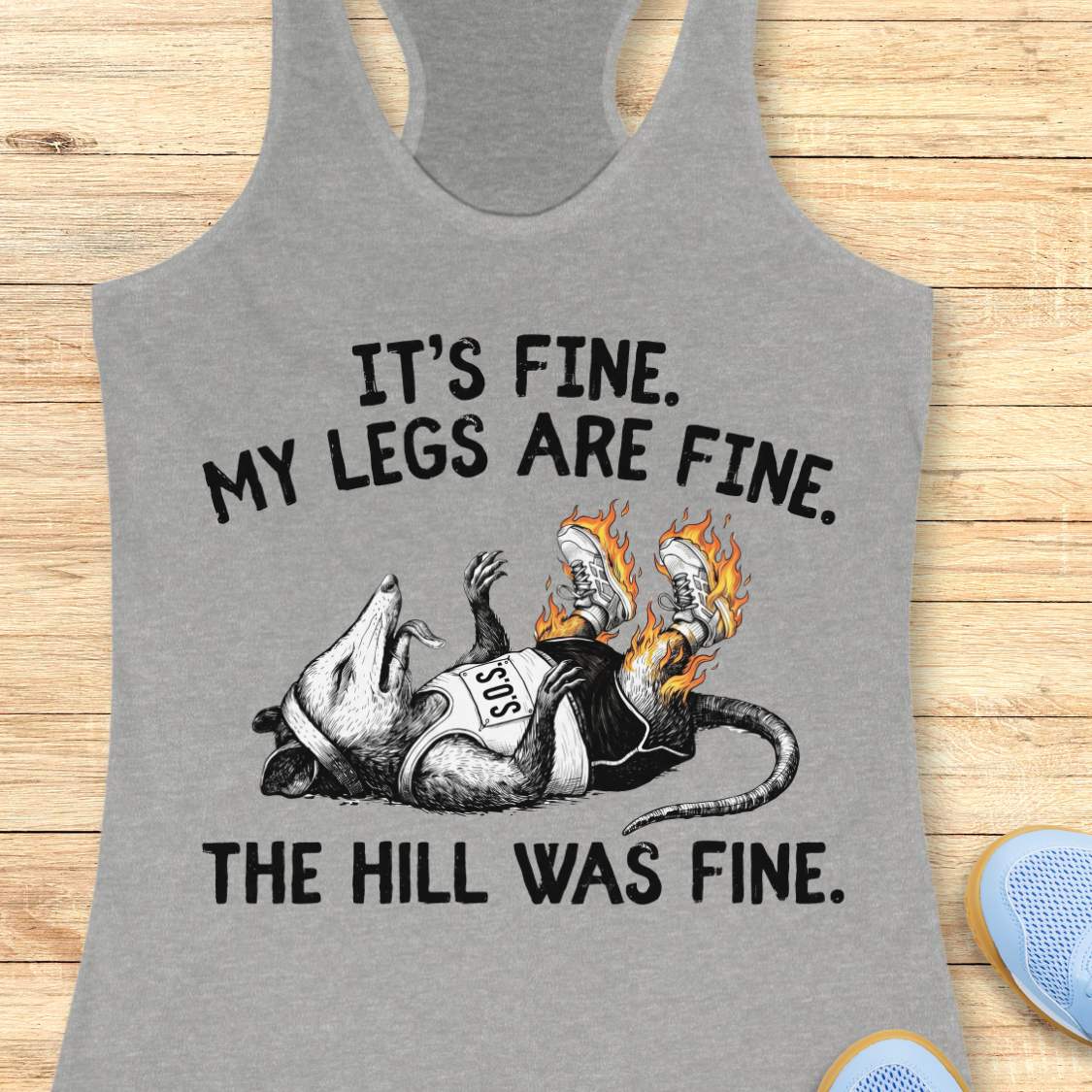 heather grey tank top with black graphic design and text 'it's fine. My legs are fine. The hill was fine.', on a wooden surface