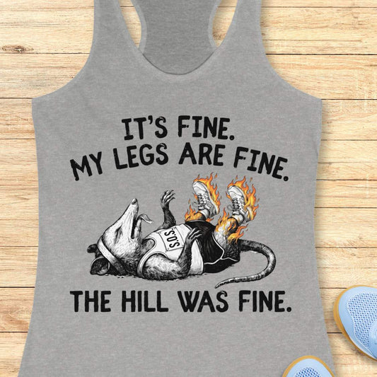 heather grey tank top with black graphic design and text 'it's fine. My legs are fine. The hill was fine.', on a wooden surface