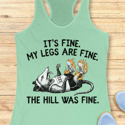mint tank top with black graphic design and text 'it's fine. My legs are fine. The hill was fine.', on a wooden surface