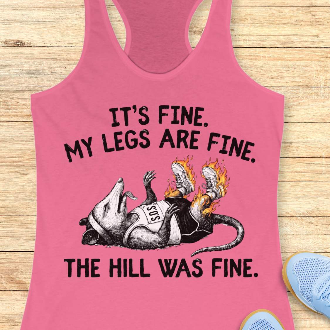 pink tank top with black graphic design and text 'it's fine. My legs are fine. The hill was fine.', on a wooden surface
