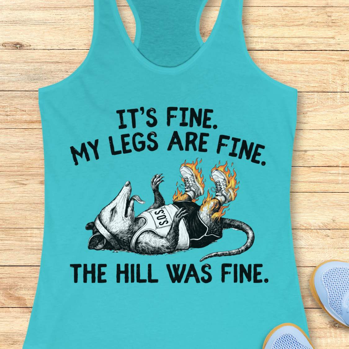blue tank top with black graphic design and text 'it's fine. My legs are fine. The hill was fine.', on a wooden surface