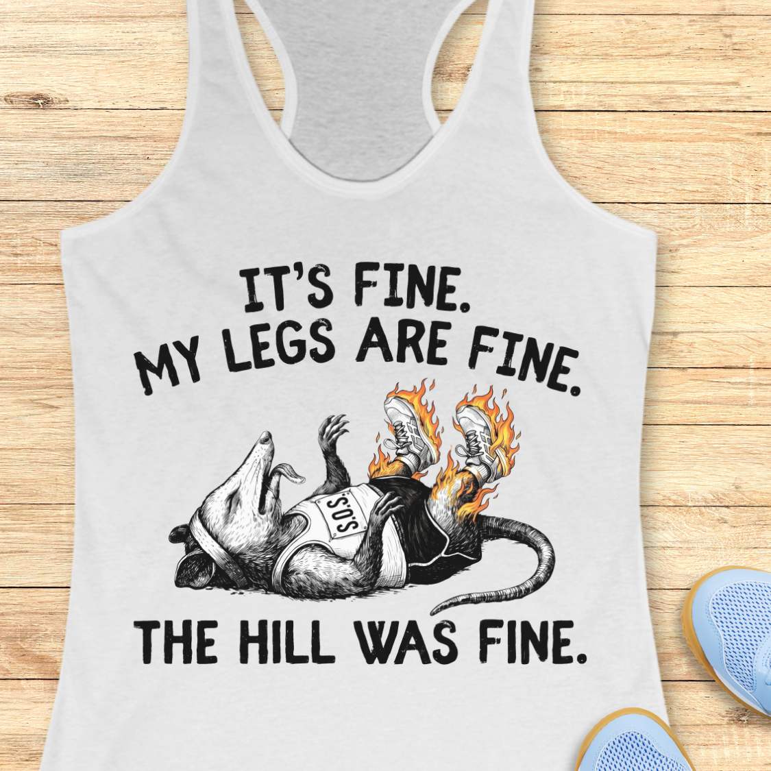 white tank top with black graphic design and text 'it's fine. My legs are fine. The hill was fine.', on a wooden surface