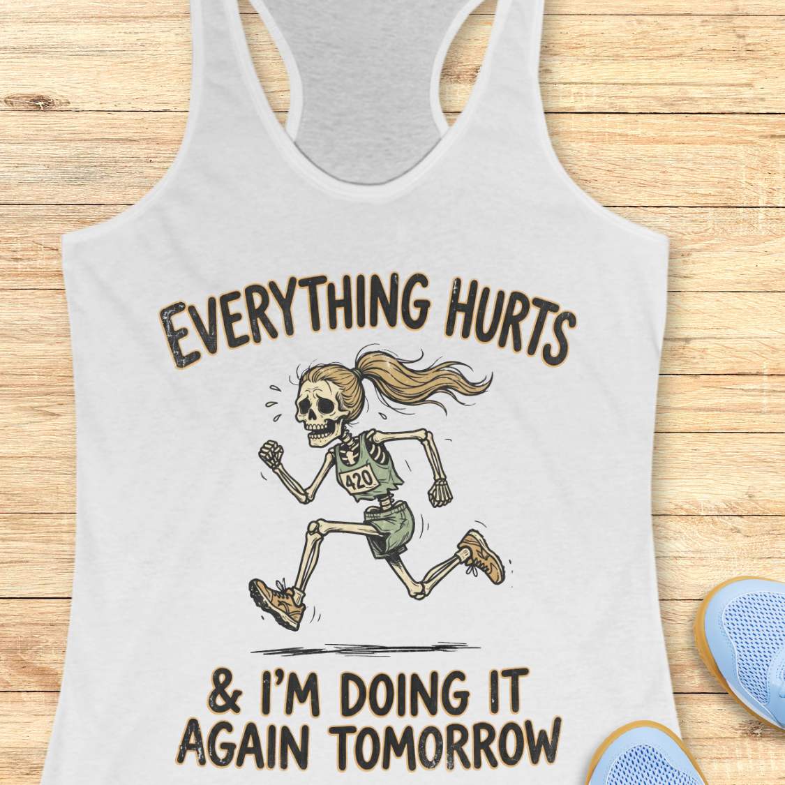 white tank top with graphic design of a runner skeleton and text 'Everything hurts and I'm doing it again tomorrow', on a wooden surface