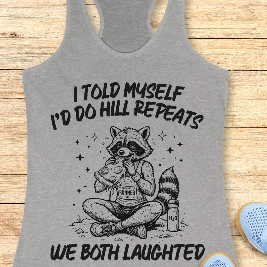 A grey racerback tank top laid flat on a light wood background, featuring a humorous text 'I told myself I'd do hill repeats. We both laughed'.