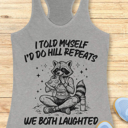 A grey racerback tank top laid flat on a light wood background, featuring a humorous text 'I told myself I'd do hill repeats. We both laughed'.