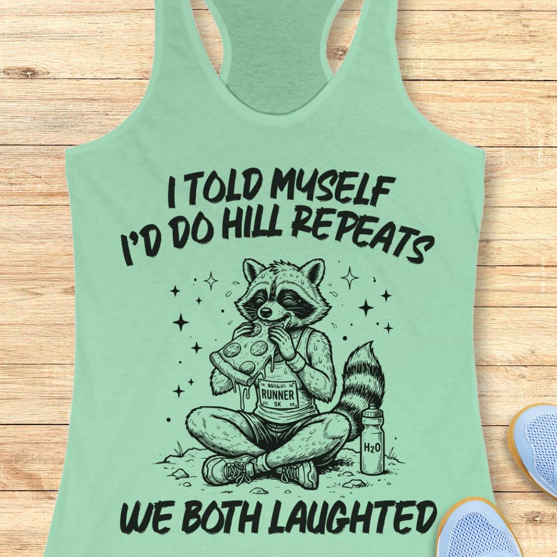A mint racerback tank top laid flat on a light wood background, featuring a humorous text 'I told myself I'd do hill repeats. We both laughed'.