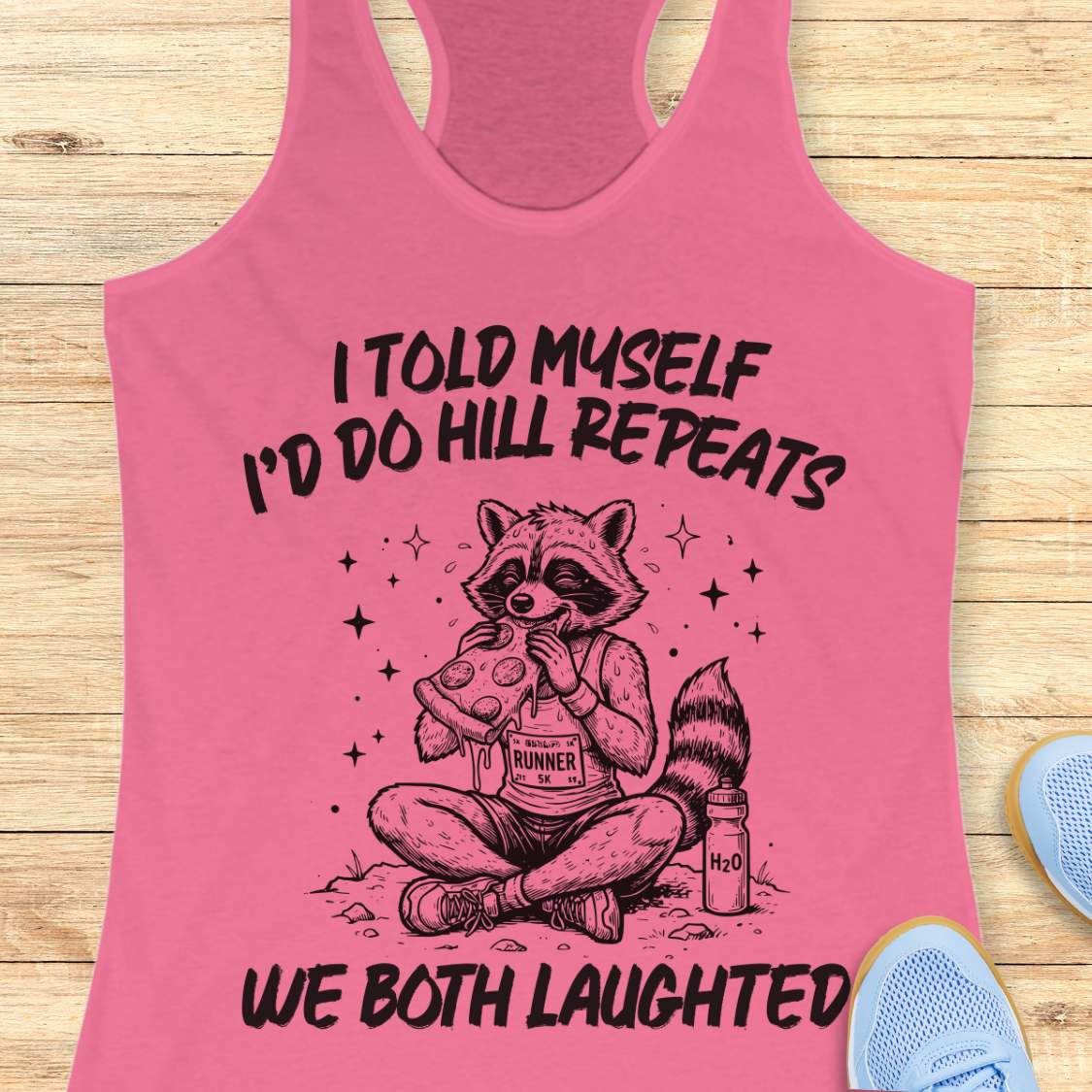 A pink racerback tank top laid flat on a light wood background, featuring a humorous text 'I told myself I'd do hill repeats. We both laughed'.