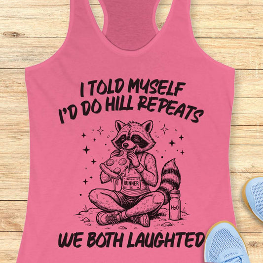 A pink racerback tank top laid flat on a light wood background, featuring a humorous text 'I told myself I'd do hill repeats. We both laughed'.