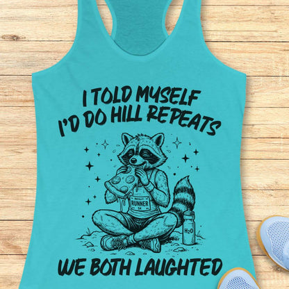 A blue racerback tank top laid flat on a light wood background, featuring a humorous text 'I told myself I'd do hill repeats. We both laughed'.