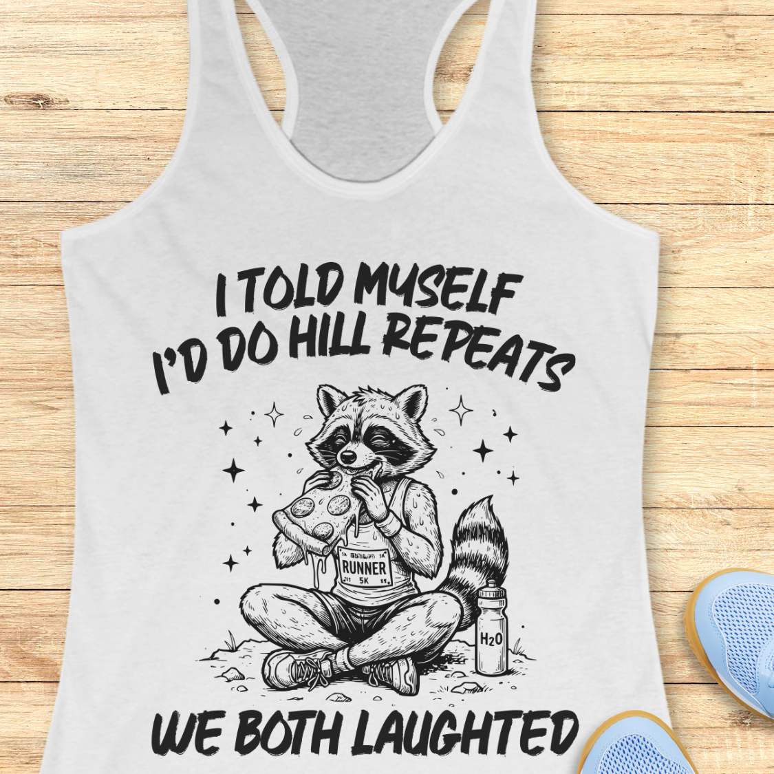 A white racerback tank top laid flat on a light wood background, featuring a humorous text 'I told myself I'd do hill repeats. We both laughed'.
