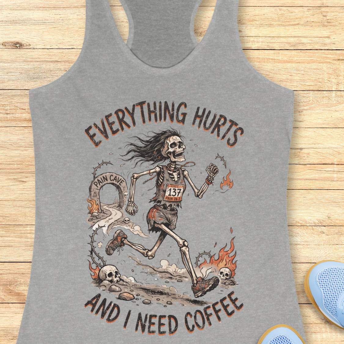 A grey racerback tank top laid flat on a light wood background, featuring a humorous skeleton runner graphic with the text 'everything hurts and I need coffee'.