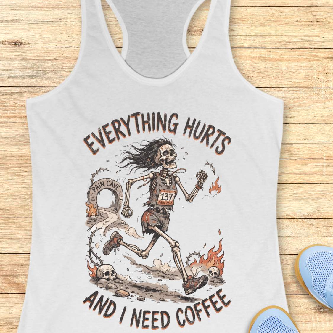 A white racerback tank top laid flat on a light wood background, featuring a humorous skeleton runner graphic with the text 'everything hurts and I need coffee'.