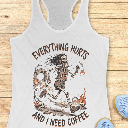 A white racerback tank top laid flat on a light wood background, featuring a humorous skeleton runner graphic with the text 'everything hurts and I need coffee'.