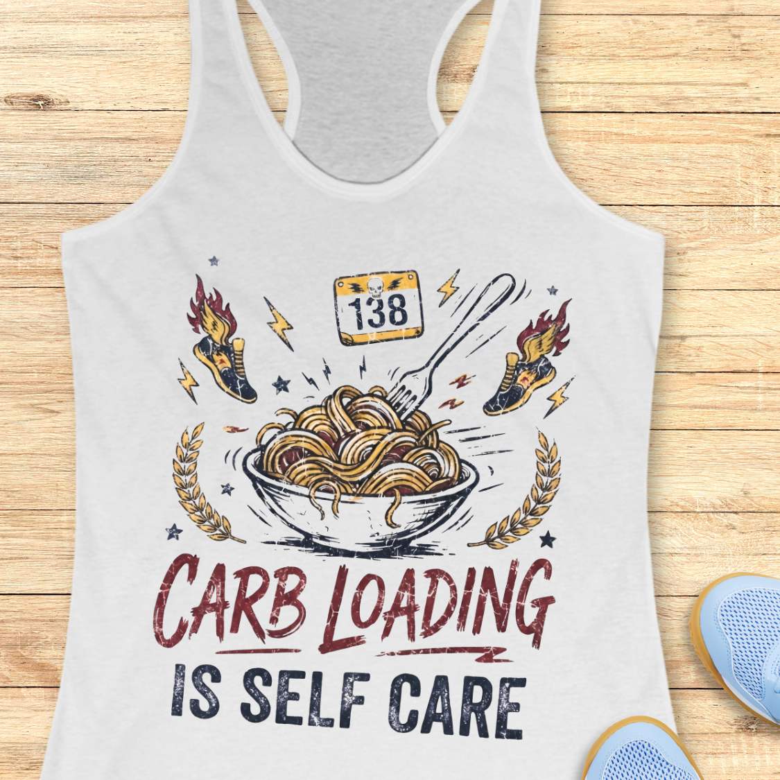 A white racerback tank top laid flat on a light wood background, featuring a bowl with noodles graphic with the text 'carb loading is self care'.