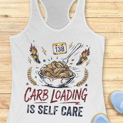 A white racerback tank top laid flat on a light wood background, featuring a bowl with noodles graphic with the text 'carb loading is self care'.