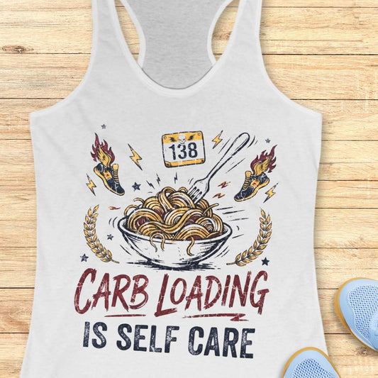 A white racerback tank top laid flat on a light wood background, featuring a bowl with noodles graphic with the text 'carb loading is self care'.