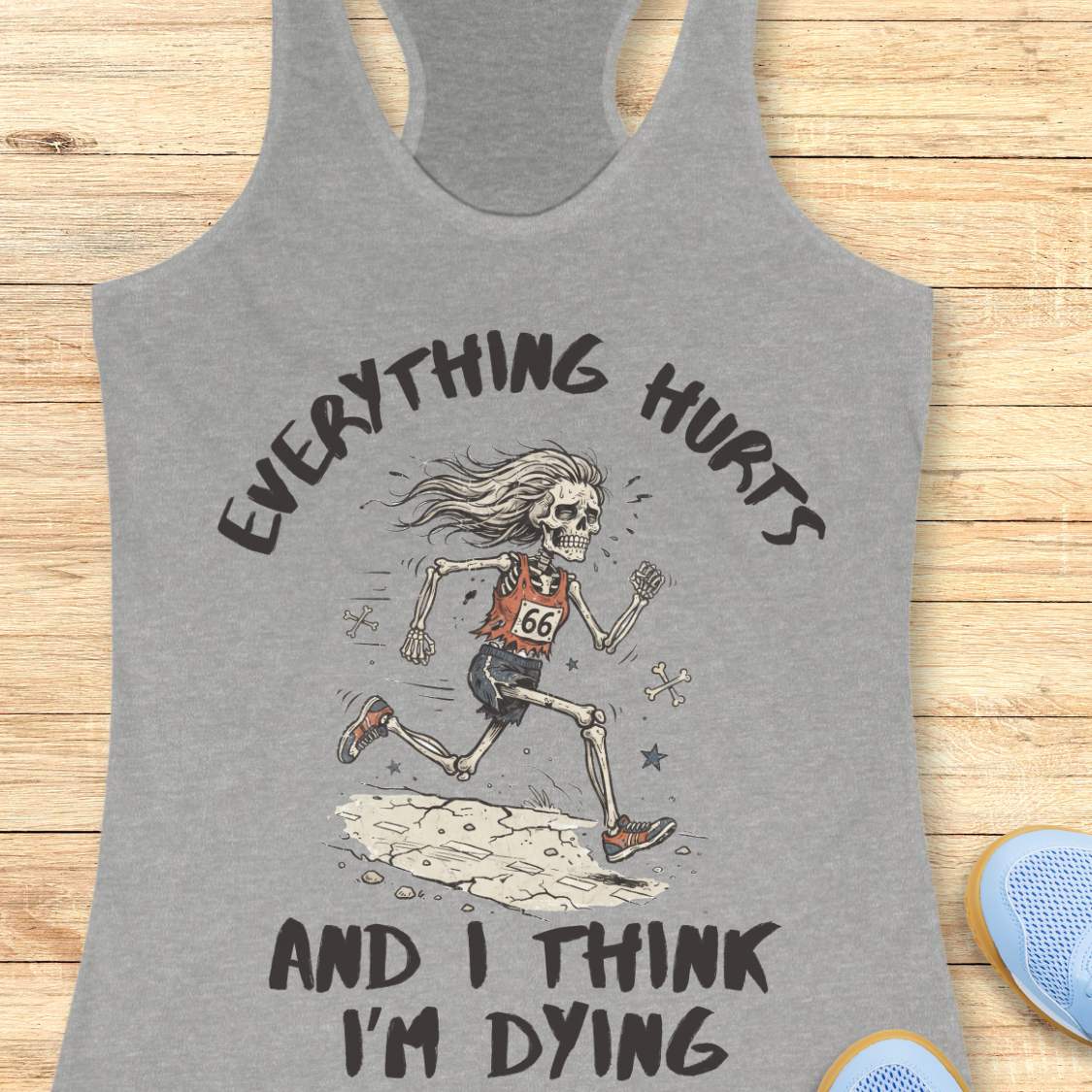 A grey racerback tank top laid flat on a light wood background, featuring a humorous skeleton runner graphic with the text 'everything hurts and I think I'm dying'.