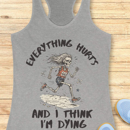 A grey racerback tank top laid flat on a light wood background, featuring a humorous skeleton runner graphic with the text 'everything hurts and I think I'm dying'.