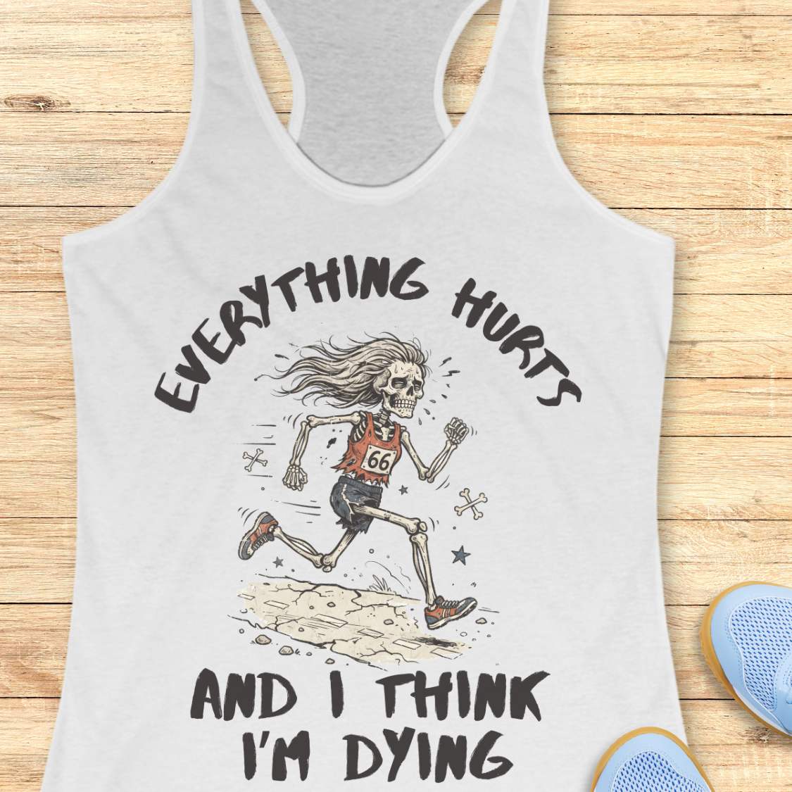 A white racerback tank top laid flat on a light wood background, featuring a humorous skeleton runner graphic with the text 'everything hurts and I think I'm dying'.