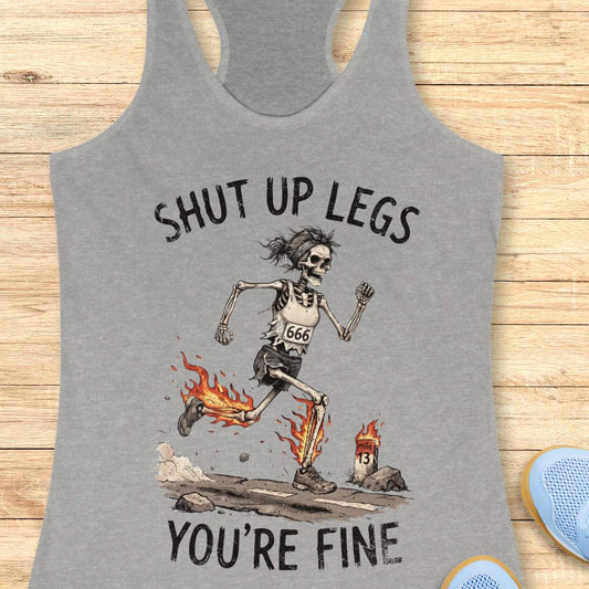 A heather gray racerback tank top laid flat on a light wood background, featuring a humorous running skeleton graphic with the text 'Shut up legs you're fine'.