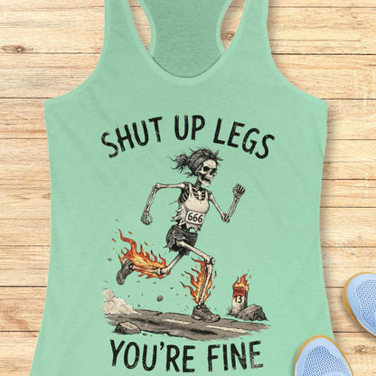 A mint racerback tank top laid flat on a light wood background, featuring a humorous running skeleton graphic with the text 'Shut up legs you're fine'.