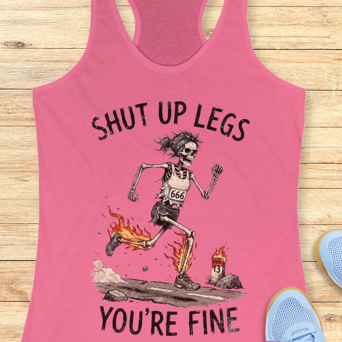 A  pink racerback tank top laid flat on a light wood background, featuring a humorous running skeleton graphic with the text 'Shut up legs you're fine'.