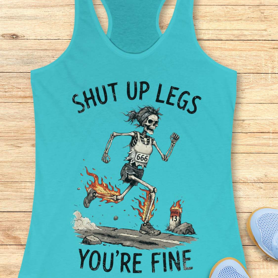 A blue racerback tank top laid flat on a light wood background, featuring a humorous running skeleton graphic with the text 'Shut up legs you're fine'.