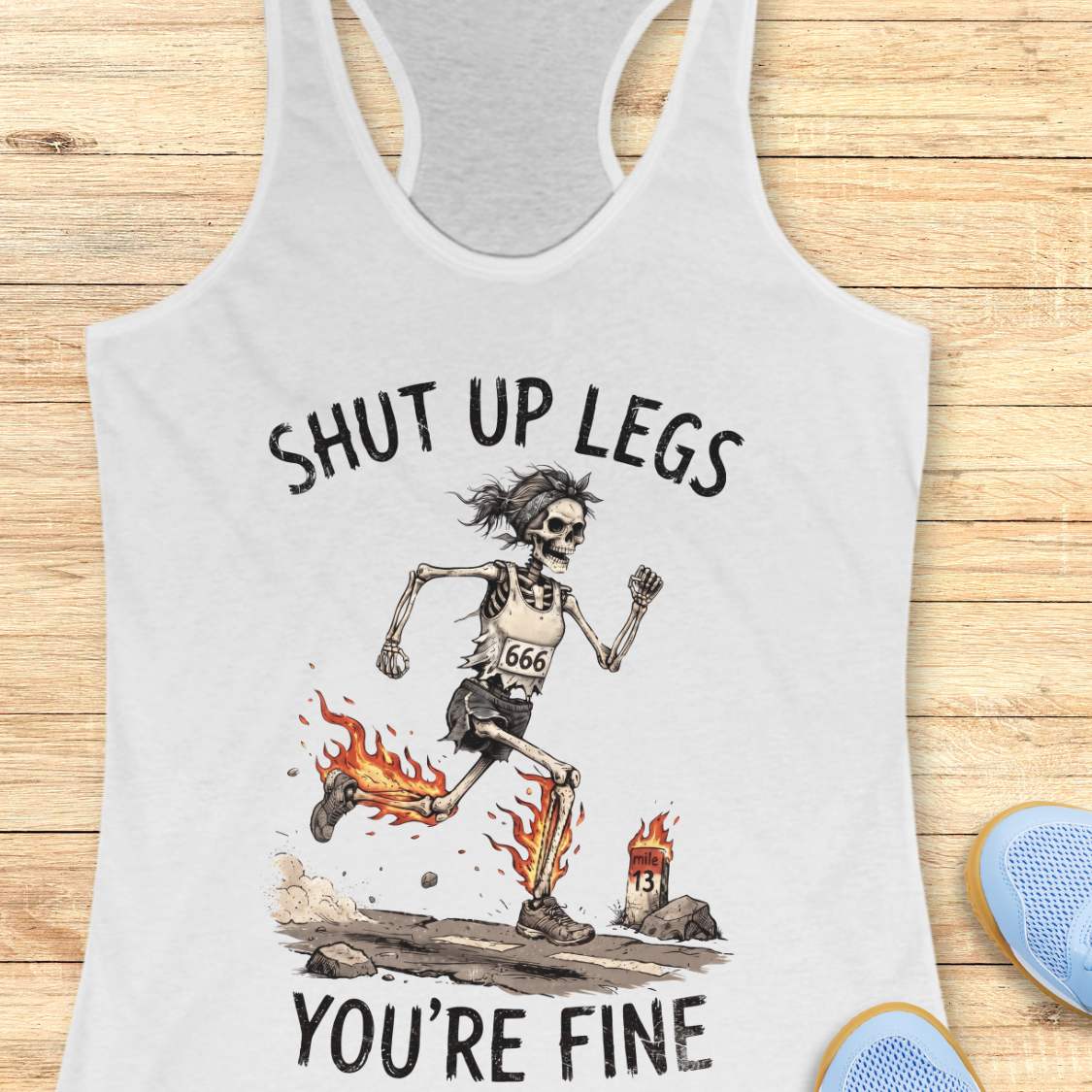 A white racerback tank top laid flat on a light wood background, featuring a humorous running skeleton graphic with the text 'Shut up legs you're fine'.