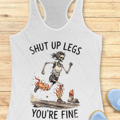 A white racerback tank top laid flat on a light wood background, featuring a humorous running skeleton graphic with the text 'Shut up legs you're fine'.