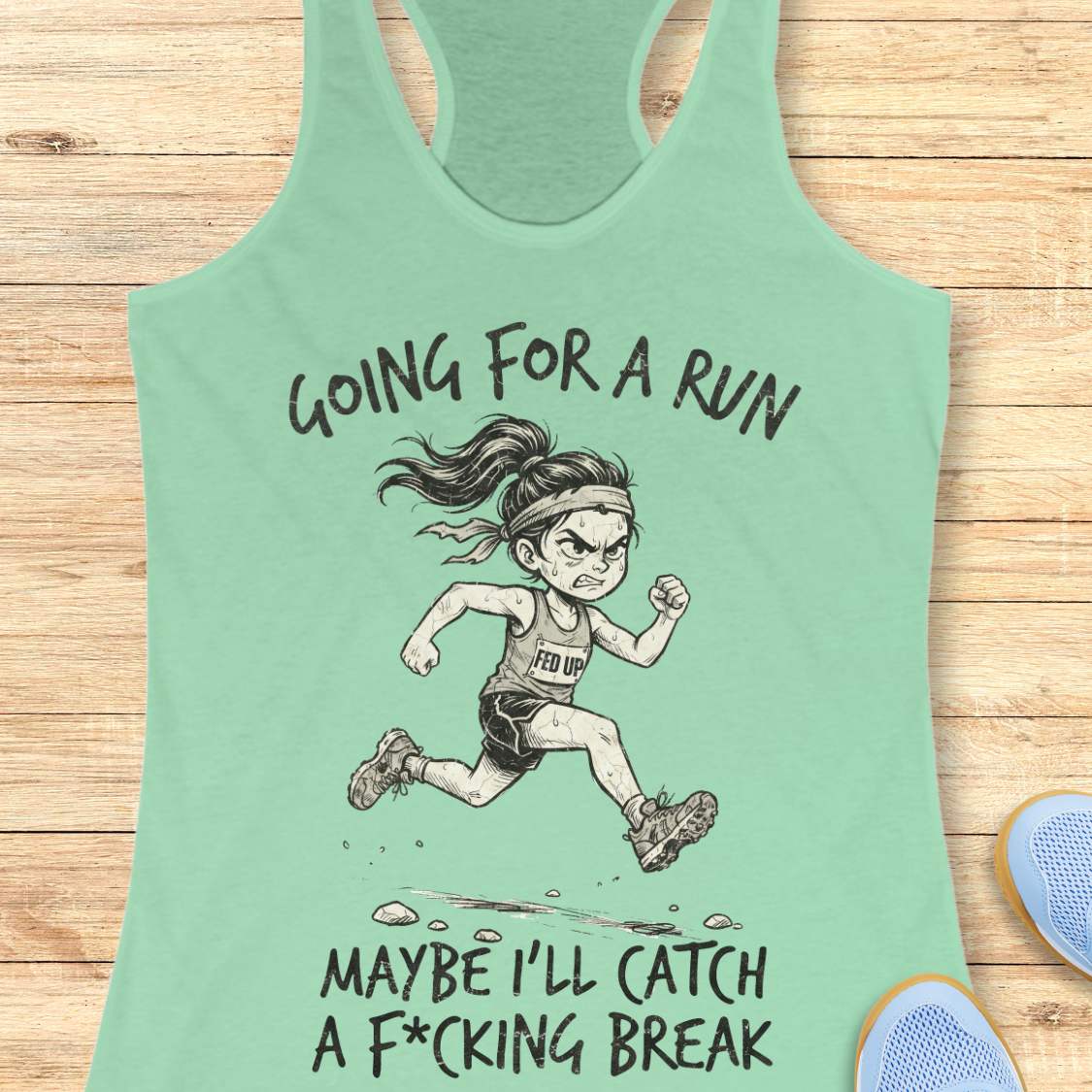 A mint green racerback tank top laid flat on a light wood background, featuring a black line art illustration of an angry female runner with the text 'GOING FOR A RUN' above and 'MAYBE I'LL CATCH A F*CKING BREAK' below.