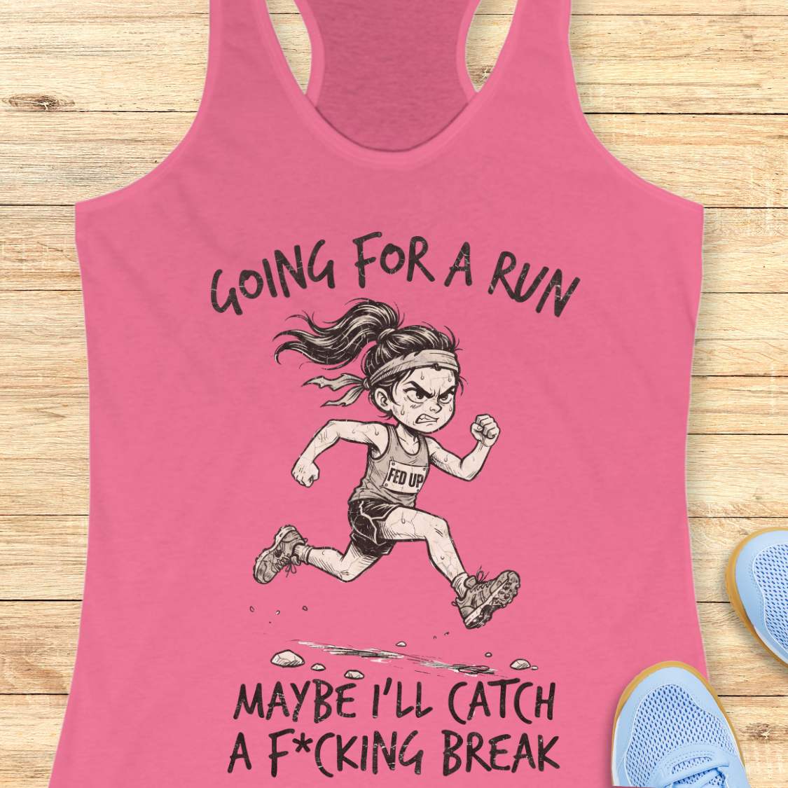 A pink racerback tank top laid flat on a light wood background, featuring a black line art illustration of an angry female runner with the text 'GOING FOR A RUN' above and 'MAYBE I'LL CATCH A F*CKING 