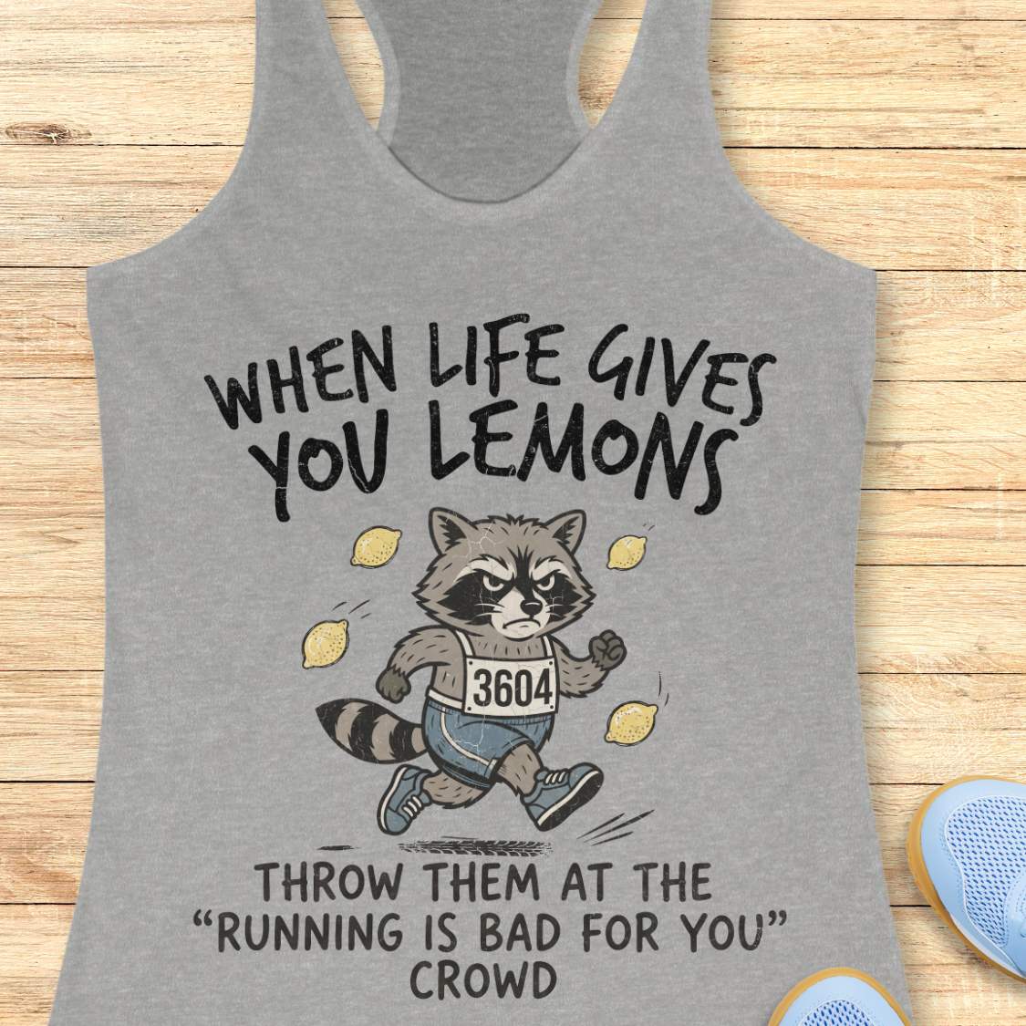 A heather gray racerback tank top laid flat on a light wood surface, featuring a printed graphic of a raccoon running with lemons and the text 'WHEN LIFE GIVES YOU LEMONS THROW THEM AT THE RUNNING IS BAD FOR YOU CROWD.'.