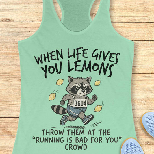 A mint green racerback tank top laid flat on a light wood background, featuring a cartoon raccoon running with lemons and the text 'WHEN LIFE GIVES YOU LEMONS' and 'THROW THEM AT THE RUNNING IS BAD FOR YOU CROWD'.