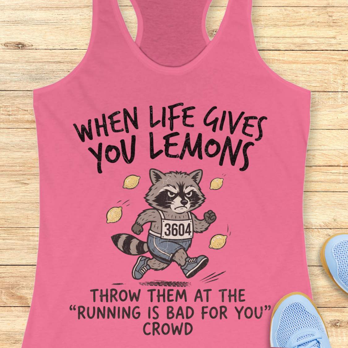 A pink racerback tank top laid flat on a light wood surface, featuring a cartoon raccoon running with the text 'WHEN LIFE GIVES YOU LEMONS' above and 'THROW THEM AT THE RUNNING IS BAD FOR YOU CROWD' below. Partial blue sneakers are visible at the edges as styling props.