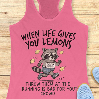 A pink racerback tank top laid flat on a light wood surface, featuring a cartoon raccoon running with the text 'WHEN LIFE GIVES YOU LEMONS' above and 'THROW THEM AT THE RUNNING IS BAD FOR YOU CROWD' below. Partial blue sneakers are visible at the edges as styling props.