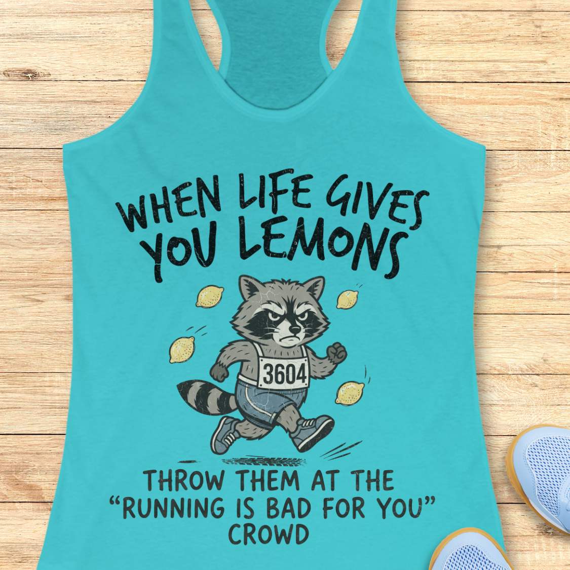 A turquoise racerback tank top laid flat on a light wood surface, featuring a humorous raccoon graphic running with lemons and the text 'WHEN LIFE GIVES YOU LEMONS THROW THEM AT THE RUNNING IS BAD FOR YOU CROWD.' A partial blue shoe is visible at the lower right edge.