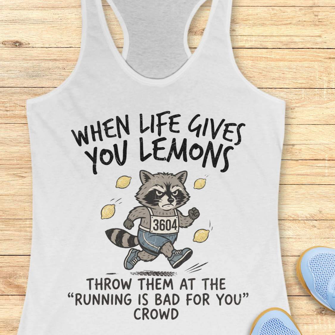 A white racerback tank top laid flat on a light wood surface, featuring a humorous raccoon graphic with the text 'WHEN LIFE GIVES YOU LEMONS THROW THEM AT THE "RUNNING IS BAD FOR YOU" CROWD'. Partial blue sneakers are visible at the bottom right as styling props.