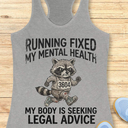 A heather gray racerback tank top laid flat on a light wood background, featuring a black graphic print of a raccoon running with the text 'RUNNING FIXED MY MENTAL HEALTH' above and 'MY BODY IS SEEKING LEGAL ADVICE' below.