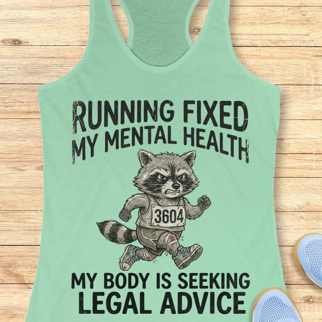 A mint green racerback tank top laid flat on a light wood background, featuring a graphic of a raccoon runner with the text 'RUNNING FIXED MY MENTAL HEALTH MY BODY IS SEEKING LEGAL ADVICE'.