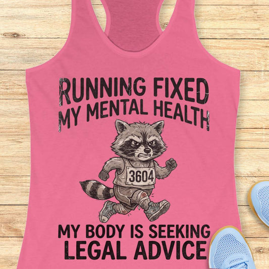 A pink racerback tank top laid flat on a light wood surface, featuring a distressed black graphic that reads 'RUNNING FIXED MY MENTAL HEALTH' above a raccoon running with a race bib '3604', and 'MY BODY IS SEEKING LEGAL ADVICE' below. Partial blue sneakers are visible at the edges as styling props.