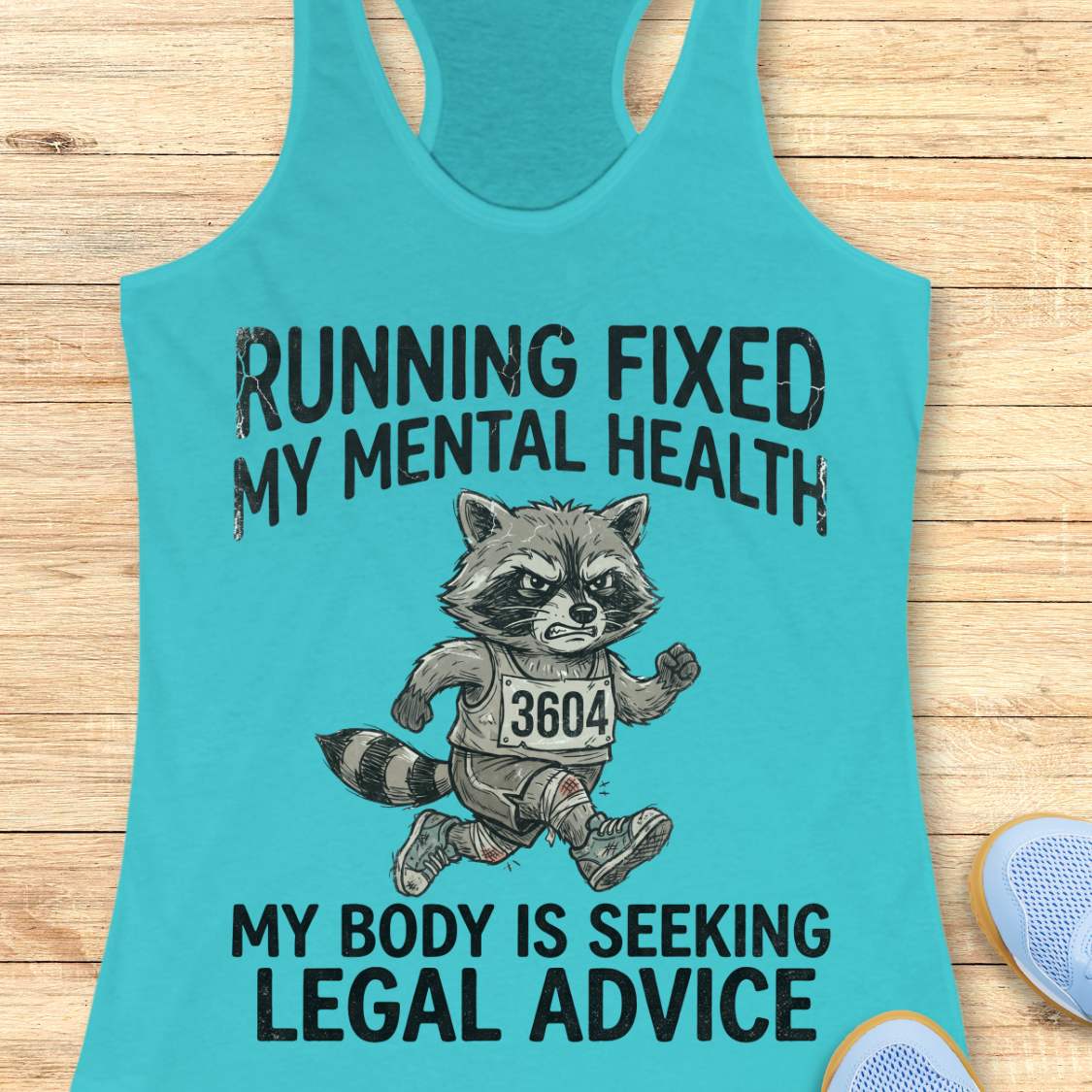 A turquoise racerback tank top laid flat on a light wood surface, featuring a black graphic of a raccoon running with the text 'RUNNING FIXED MY MENTAL HEALTH MY BODY IS SEEKING LEGAL ADVICE'. A partial light blue shoe is visible at the bottom right.