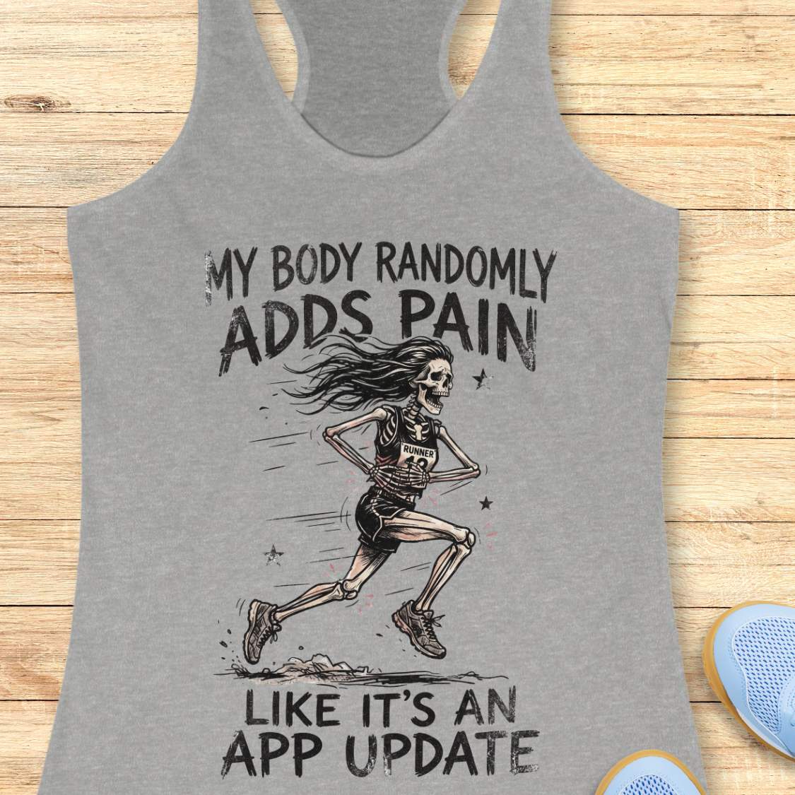 A heather gray racerback tank top laid flat on a light wood surface, featuring a black graphic of a running skeleton and the text 'MY BODY RANDOMLY ADDS PAIN LIKE IT'S AN APP UPDATE'. Partial blue shoes are visible at the bottom right as styling props.