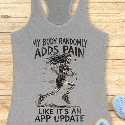 A heather gray racerback tank top laid flat on a light wood surface, featuring a black graphic of a running skeleton and the text 'MY BODY RANDOMLY ADDS PAIN LIKE IT'S AN APP UPDATE'. Partial blue shoes are visible at the bottom right as styling props.