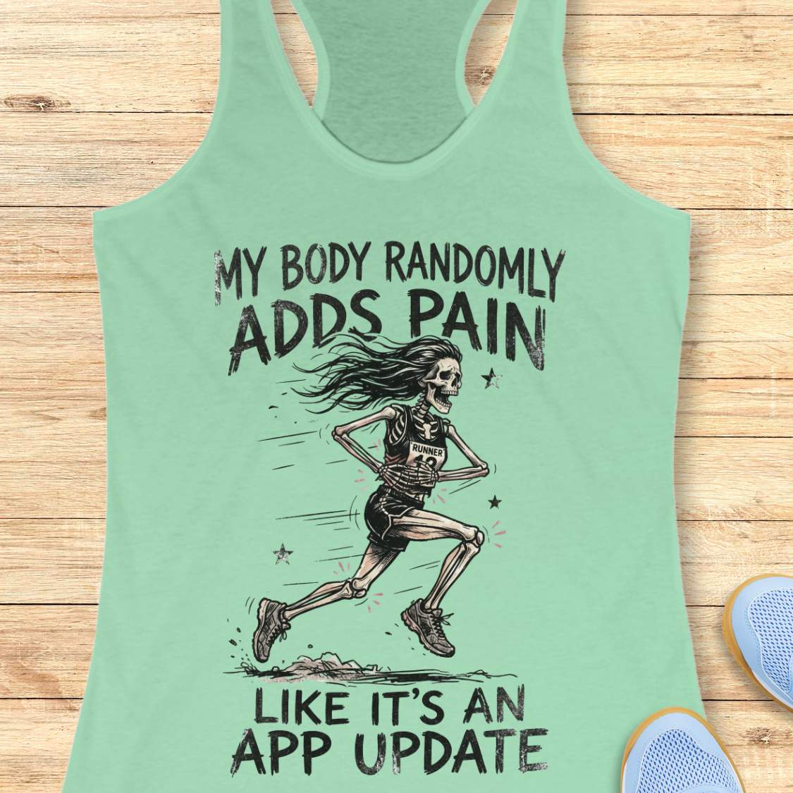 A mint green racerback tank top laid flat on a light wood background, featuring a black graphic of a running skeleton and the text 'MY BODY RANDOMLY ADDS PAIN LIKE IT'S AN APP UPDATE'. Partial blue shoes are visible at the bottom right as styling props.