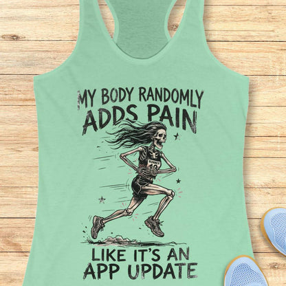 A mint green racerback tank top laid flat on a light wood background, featuring a black graphic of a running skeleton and the text 'MY BODY RANDOMLY ADDS PAIN LIKE IT'S AN APP UPDATE'. Partial blue shoes are visible at the bottom right as styling props.