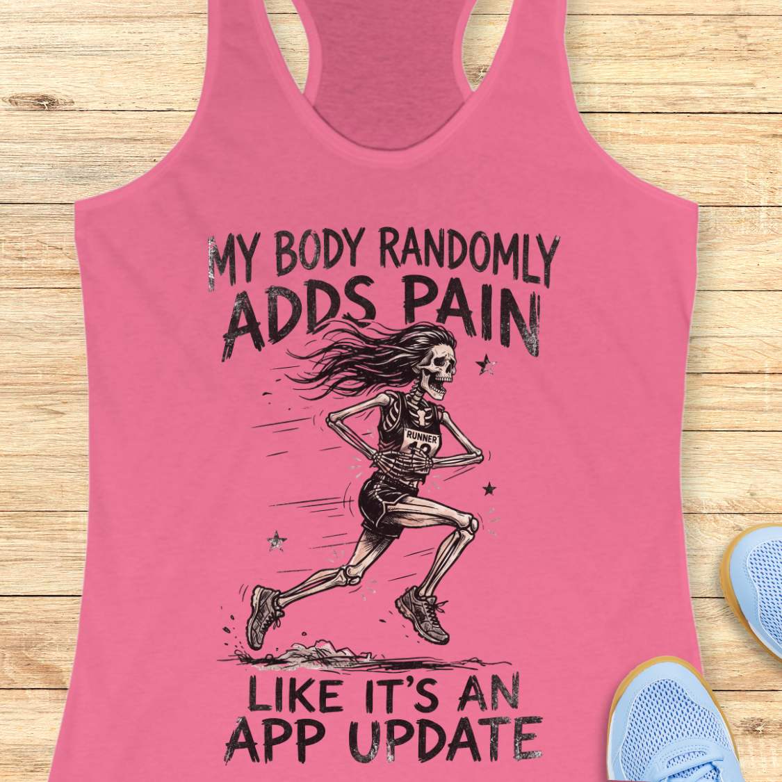 A pink racerback tank top laid flat on a light wood surface, featuring a black graphic of a running skeleton with the text 'MY BODY RANDOMLY ADDS PAIN LIKE IT'S AN APP UPDATE.' Partial blue shoes are visible at the edges as styling props.
