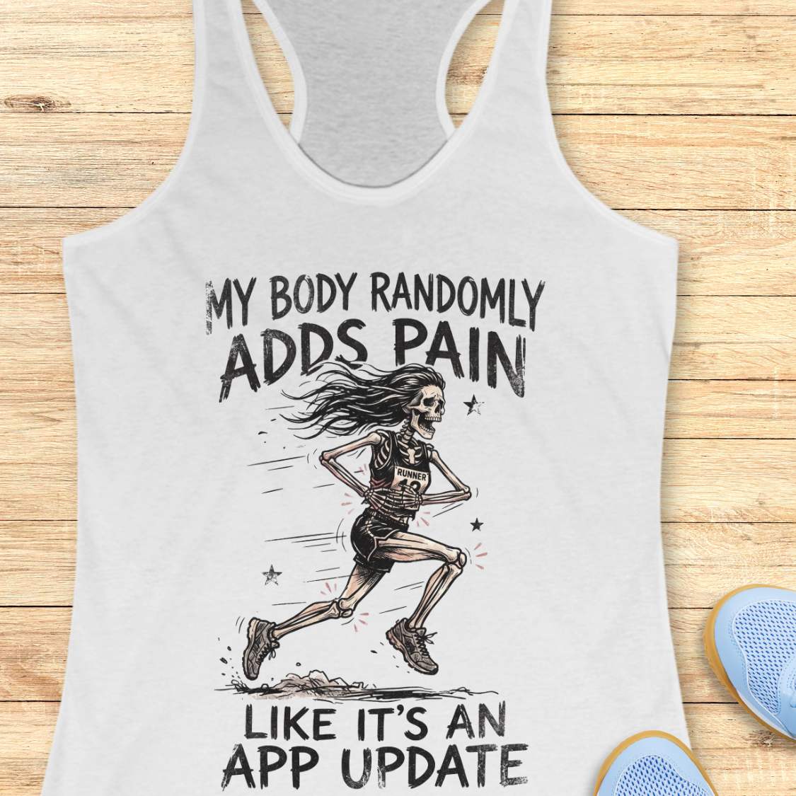 A white racerback tank top laid flat on a light wood background, featuring a black graphic of a running skeleton with the text 'MY BODY RANDOMLY ADDS PAIN LIKE IT'S AN APP UPDATE'. Partial blue shoes are visible at the bottom right as styling props.