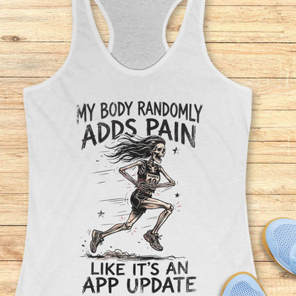 A white racerback tank top laid flat on a light wood background, featuring a black graphic of a running skeleton with the text 'MY BODY RANDOMLY ADDS PAIN LIKE IT'S AN APP UPDATE'. Partial blue shoes are visible at the bottom right as styling props.